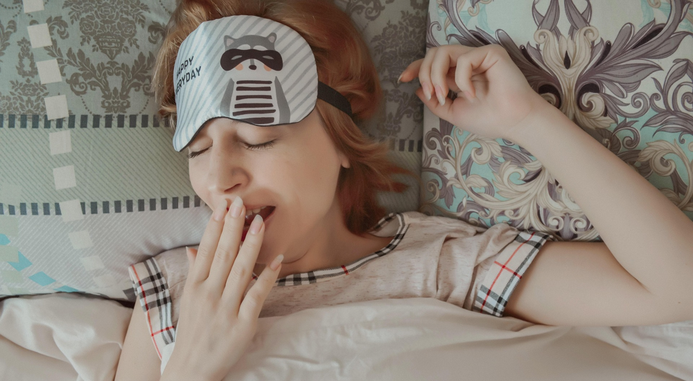 Álmos nő ásít egy ágyban fekve, alvómaszkkal a homlokán